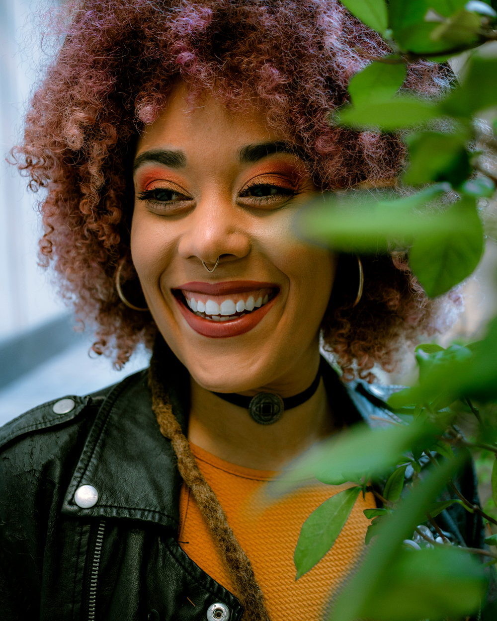 A smiling woman with a curly afro among green leaves